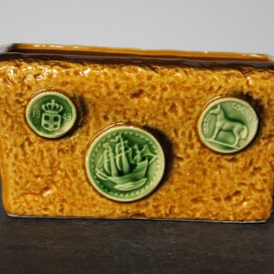 May include: A rectangular ceramic planter with a brown glaze and green accents. The planter has three decorative medallions with images of a ship, a crown, and a horse. The medallions have the text "1949" and "Cordoba" on them.