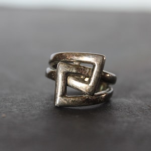 May include: A silver-toned ring with a geometric design. The ring features a square-shaped design with open spaces, creating an interesting visual effect. The ring is likely made of metal and has a slightly aged appearance.