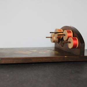 May include: A wooden spool holder with a curved top and a flat base. The holder has six spools of thread, some with red thread and some with beige thread.