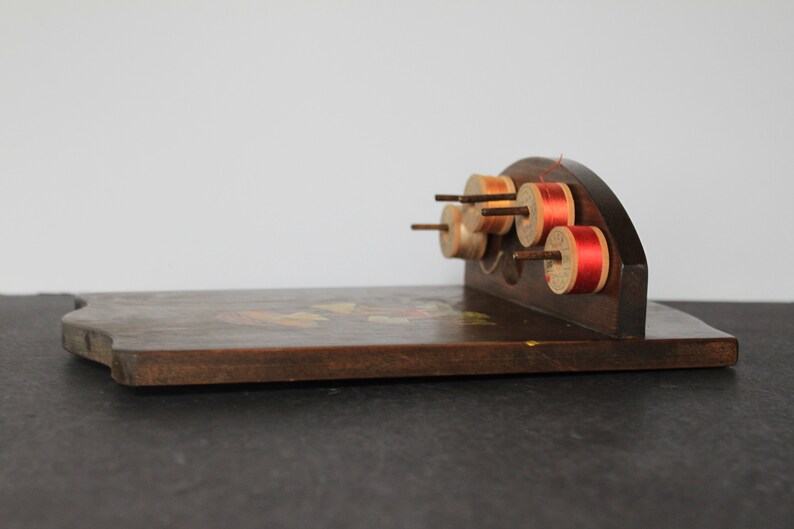 May include: A wooden spool holder with a curved top and a flat base. The holder has six spools of thread, some with red thread and some with beige thread.