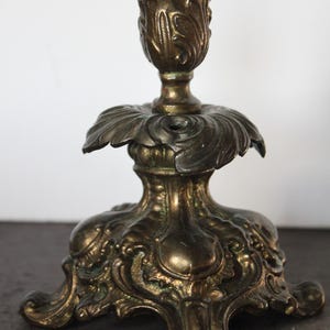 May include: Ornate brass candlestick holder with a detailed floral design. The candlestick features a wide base, a central stem, and a cup-shaped top. The brass has an aged patina, giving it a vintage appearance. A decorative piece for home decor.