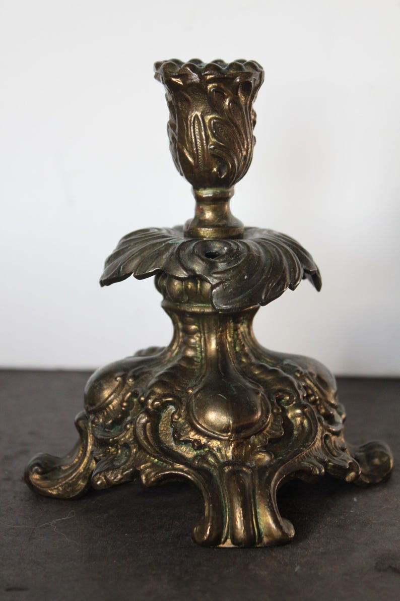 May include: Ornate brass candlestick holder with a detailed floral design. The candlestick features a wide base, a central stem, and a cup-shaped top. The brass has an aged patina, giving it a vintage appearance. A decorative piece for home decor.