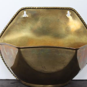 May include: Vintage brass wall pocket with a hexagonal shape and a decorative rope-like trim. The interior has a curved design, and the piece has two holes for hanging. The brass has a warm, aged patina.