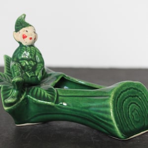 Puede incluir: Una maceta de cerámica vintage con forma de tronco con una figura de elfo sentado. La maceta es de color verde brillante con un diseño detallado de vetas de madera. El elfo tiene un atuendo verde y una cara pintada de rojo.