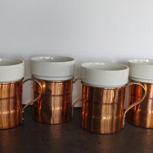 May include: Four copper mugs with white ceramic tops and gold-colored handles are arranged in a row. The copper bodies have a shiny, reflective surface. The mugs are designed with a classic, elegant aesthetic, suitable for serving beverages.