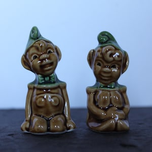 May include: Two ceramic figurines of whimsical characters. Each figure is brown with a green hat and bow tie. One figure is seated with its hands folded, while the other is kneeling with its hands on its knees. The figurines have a glossy finish.