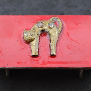 May include: A rectangular belt buckle featuring a glossy red enamel finish. Centered on the surface is a gold-toned metal cat silhouette in an arched pose, embellished with two clear, sparkling rhinestones set into the cat's back and head.