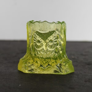May include: A lime green glass owl figurine. The owl is carved into a tree stump shape. The owl's face is visible in the center of the stump. The glass has a textured, rough appearance. The figurine is sitting on a dark surface.