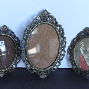 May include: Three antique oval picture frames with ornate, filigree-style metalwork. The frames are in varying sizes and feature a tarnished gold color. One frame contains a print of a couple, while the others are empty.