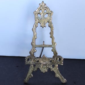 May include: Ornate brass easel with a decorative floral design. The small display stand has a three-legged base and a hinged back support. Ideal for displaying small artwork or photos.