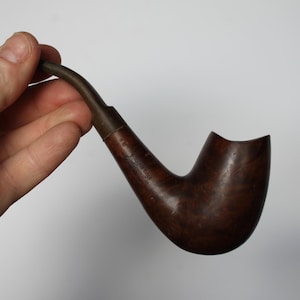 May include: A brown wooden pipe with a curved stem and a bowl. The pipe is held in a hand against a white background. The pipe appears to be made of a dark wood with a smooth finish.
