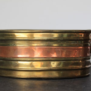 May include: Oval brass planter with a copper band around the middle. The planter has two handles on the sides.