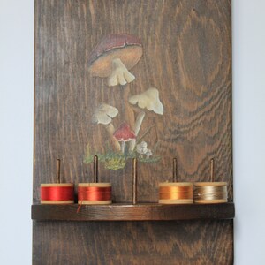 May include: Wooden wall-mounted thread rack with a painted mushroom design. The rack has five spools of thread, two red, one orange, and two beige.