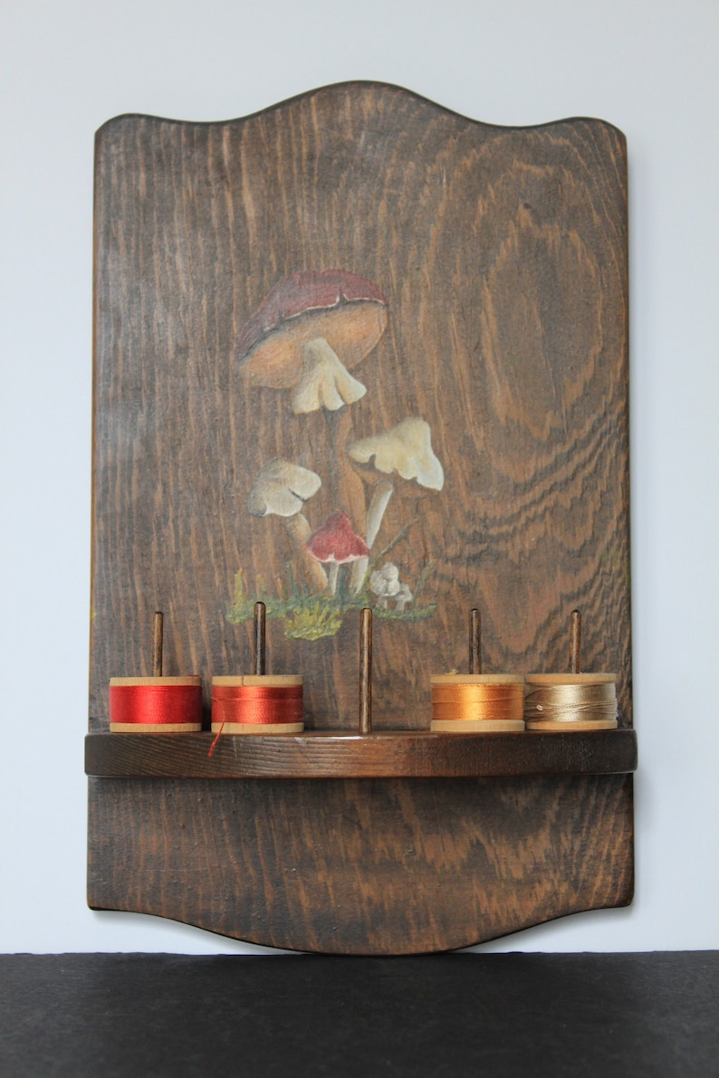 May include: Wooden wall-mounted thread rack with a painted mushroom design. The rack has five spools of thread, two red, one orange, and two beige.