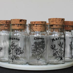 May include: A set of clear glass spice jars with cork lids, arranged on a white circular tray. Each jar is labeled with a black botanical illustration and the name of a spice, including "oregano" and "cloves".