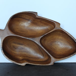 Puede incluir: Una bandeja de servir de madera con forma de hoja con tres compartimentos. La bandeja está hecha de madera de color marrón oscuro con un patrón de vetas natural. Los compartimentos tienen forma ovalada y parecen estar tallados en la madera.