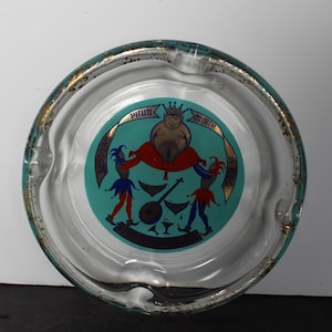 May include: Clear glass ashtray with a teal center featuring a whimsical design. The design includes a crowned figure, two jesters, and the words "PHUN MIRTH". The ashtray has a gold-colored rim and is likely vintage.