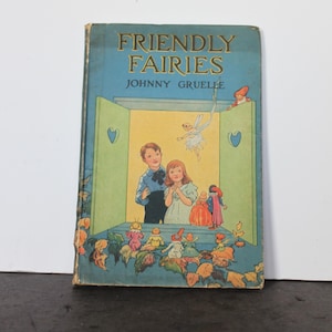 May include: A vintage book titled "Friendly Fairies" by Johnny Gruelle. The cover features a blue background with illustrations of children and fairies. The book has a worn appearance, suggesting it is an antique.