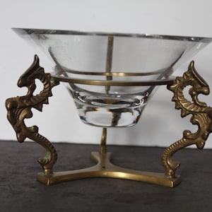 May include: A clear glass bowl with a decorative brass stand. The stand features ornate, dragon-like figures on either side and a three-pronged base. The bowl is supported by a brass ring and a central stem.