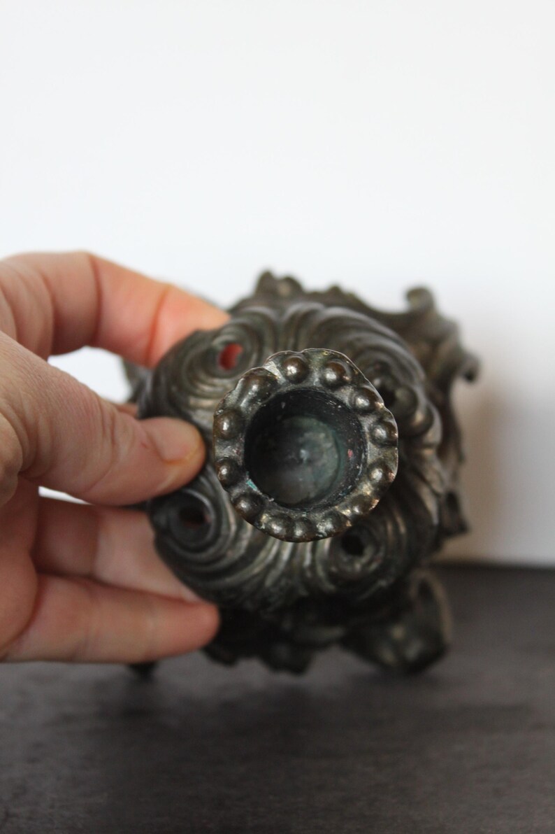 May include: Ornate, antique bronze candlestick holder with a detailed, swirling design. The candle holder has a circular opening and a decorative, textured surface. The overall design is reminiscent of a vintage, classical style.