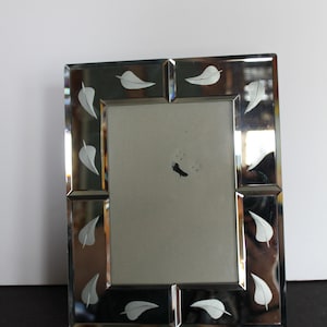 May include: A rectangular mirrored picture frame with a silver finish. The frame is decorated with white leaf designs. The frame has a neutral-colored backing and is suitable for displaying photographs or artwork.
