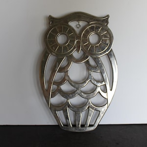 May include: Silver-colored owl-shaped trivet. The trivet is made of metal and has a cutout design. The owl's eyes are circular with radiating lines, and the body features a scalloped pattern. A small loop is at the top for hanging. The trivet is approximately 7 inches tall.