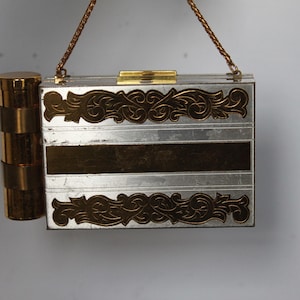 May include: A rectangular silver and gold metal clutch bag featuring an attached cylindrical gold lipstick case on the side. The clutch has a horizontal gold band across the center and ornate gold scrollwork patterns on the top and bottom edges.