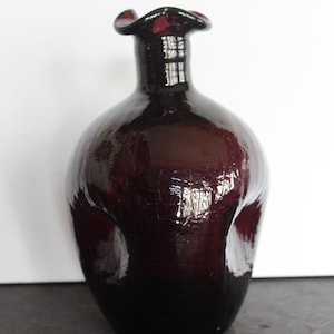 May include: A dark burgundy-colored decorative glass vase with a bulbous body and a flared, ruffled rim. The vase has a glossy finish and a textured surface, reflecting light. The design is a unique, artistic piece, suitable for home decor.