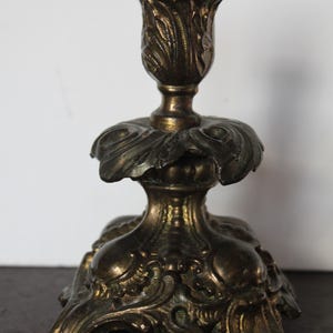 May include: Ornate antique brass candlestick with a floral design. The candlestick features a detailed, textured surface with a tulip-shaped top and a wide, decorative base. The overall design is vintage and elegant.