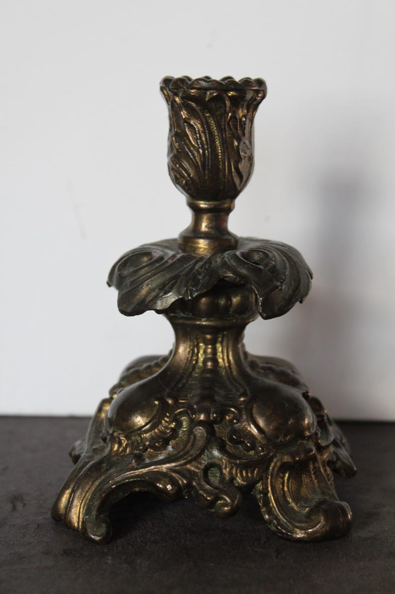 May include: Ornate antique brass candlestick with a floral design. The candlestick features a detailed, textured surface with a tulip-shaped top and a wide, decorative base. The overall design is vintage and elegant.
