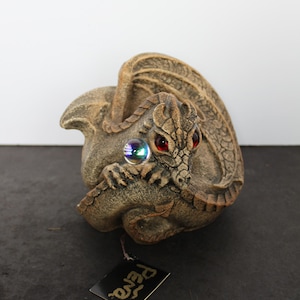 May include: A stone-textured dragon figurine in a curled position with detailed scales and wings. The dragon has bright red eyes and holds a small, iridescent spherical orb. A black square tag with the gold text Pena is attached to the base.