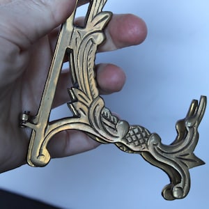 May include: Ornate brass picture frame stand with a decorative floral design. The stand is in the shape of the letter "A" and features intricate detailing. The stand is held by a hand against a white background.