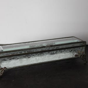 May include: A rectangular glass trinket box with a clear, textured body and a hinged lid. The box has a dark metal frame and decorative feet. The glass has a frosted appearance, and the box is set against a dark surface.