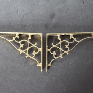 May include: Two ornate, gold-colored metal shelf brackets. Each bracket has a triangular shape with a decorative, swirling pattern. The brackets are displayed against a dark background.
