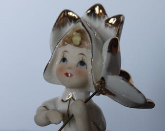 May include: A vintage porcelain figurine of a girl in a white dress and a flower-shaped hat, accented with gold trim. The figurine holds a flower stem, with delicate facial features and blue eyes. A charming collectible.