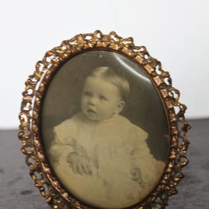 May include: Antique oval picture frame with a sepia-toned photograph of a baby. The frame is made of ornate, gold-colored metal with a detailed, filigree design. The baby is wearing a white dress.