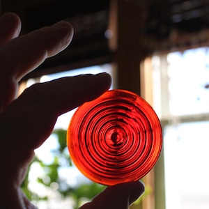 May include: A red circular lens with concentric rings. The lens is being held by a hand.