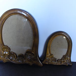 May include: Two antique, arched picture frames. The frames are a warm, golden brown color with floral detailing at the base. The oval openings are covered with a light tan fabric. One frame is larger than the other.