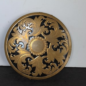 May include: A round compact mirror featuring a gold-toned metal surface with an intricate embossed acanthus leaf pattern surrounding a central circular medallion, all set against a contrasting black background within a circular gold-toned frame.