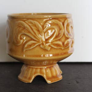 May include: A ceramic planter with a pedestal base. The planter is a warm, golden-brown color and features a raised floral design around the exterior. The base has a unique, three-footed design. This decorative piece is suitable for small plants.