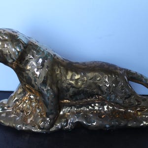 May include: A brass-colored sculpture of a roaring feline, possibly a lion or tiger, in a reclining pose. The sculpture has a textured surface and is set against a blue background. The artwork is a decorative item.