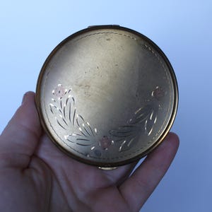 May include: A vintage, round, gold-toned compact. The compact features an etched floral design with pink accents. The compact is held in a hand against a light blue background. The compact has a hinged opening and a clasp.