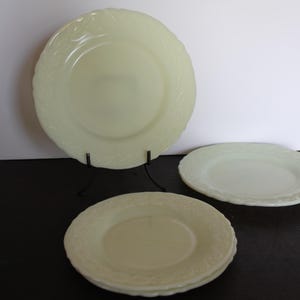 May include: Three white milk glass plates with a raised floral design. The plates are different sizes, with the largest plate on a black stand.