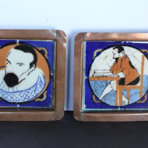 May include: Two square ceramic tiles with copper frames. Each tile features a portrait within a blue border. One depicts a man with a black beard, the other a person seated at a desk. The portraits are set against orange and white backgrounds.