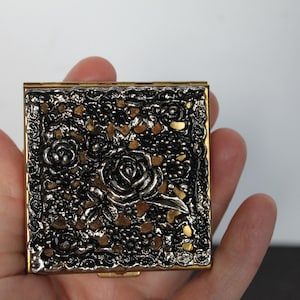 May include: A square compact mirror case featuring a metallic gold frame and a dark, textured top surface with a raised silver-toned floral rose pattern. The intricate design includes detailed petals and leaves set against a dark, openwork background.