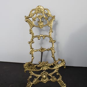 May include: A gold-colored metal display stand featuring an ornate, openwork design with scrolling leaf motifs and a central shell-like crest at the top. The stand has a wide base with decorative feet and a hinged support arm on the back.