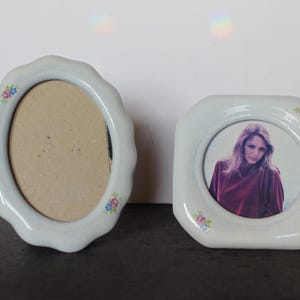 May include: Two vintage ceramic photo frames. One is oval with a scalloped edge, the other is octagonal. Both are light blue with floral accents. The octagonal frame holds a photo of a person.