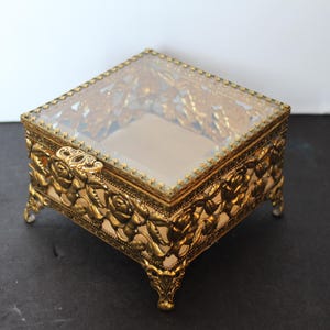 May include: A vintage gold-toned jewelry box with a clear glass top and ornate floral detailing. The box is square, with four small feet and a hinged lid. The interior is lined with a light-colored fabric.