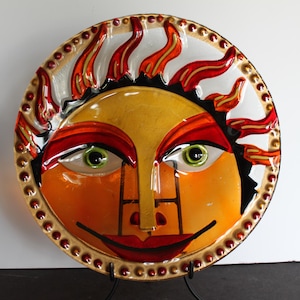 May include: A decorative ceramic plate featuring a sun face design. The sun has a yellow face, orange cheeks, red lips, and green eyes. The rim is adorned with red and gold accents, and the sun's rays are red and orange.