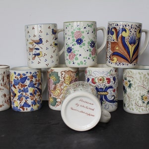May include: Set of nine ceramic mugs with various floral and bird designs. The mugs are white with blue, green, yellow, and brown accents. The bottom of one mug reads "Sarreguemines by Nicholas Wood".